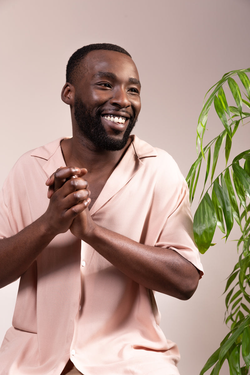 dunkelhäutiger mann lacht zur linken seite mit einem rosa farbe hemd hände zusammen fröhlich rechte seite pflanze hintergrund backdrop altrosa